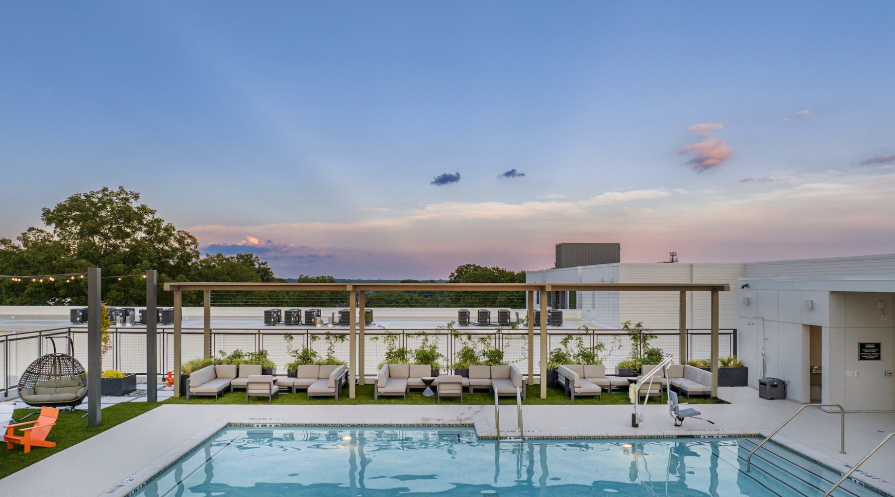 A rooftop swimming pool area at sunset, featuring a modern design with lounge chairs, cushioned seating, and a wooden pergola adorned with greenery. The pool has built-in steps and a handrail for easy access. A hanging egg chair and Adirondack chairs add 